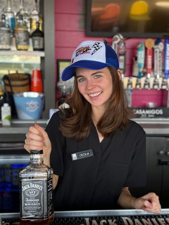 Smiling bartender in a blue trucker hat and black uniform holding a whiskey bottle behind a colorful bar counter with multiple beer taps