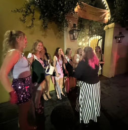 Group of women wearing sashes laughing and posing outside a lit arched entrance with lanterns on a lively downtown sidewalk at night — bachelorette night out.
