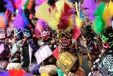 Playful row of colorful voodoo souvenir dolls with painted faces, bright feather headdresses, beads and fabric details, each with a handwritten tag on a market display