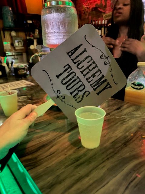 Hand holding an "Alchemy Tours" paddle above a plastic drink cup on a neon-lit marble bar counter, jar dispenser and server blurred in background