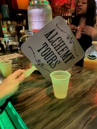 Hand holding an "Alchemy Tours" paddle above a plastic drink cup on a neon-lit marble bar counter, jar dispenser and server blurred in background