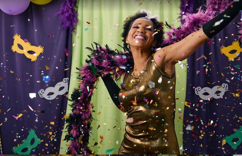 Mardi Gras–style party scene: smiling person in a gold dress and purple feather boa dancing through colorful confetti with mask decorations and purple-green-gold backdrop.
