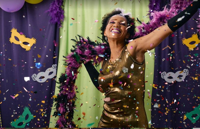 Mardi Gras–style party scene: smiling person in a gold dress and purple feather boa dancing through colorful confetti with mask decorations and purple-green-gold backdrop.