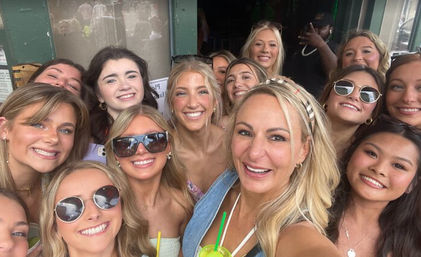 Group selfie of smiling women on a sunny outdoor patio, posing closely with sunglasses and colorful drinks for a cheerful summer gathering.