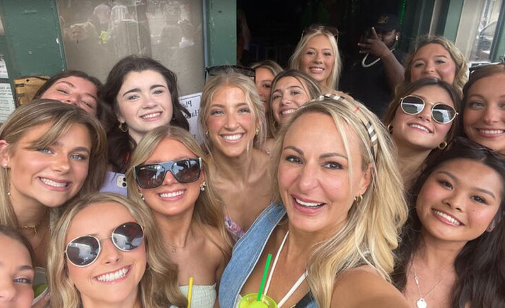 Group selfie of smiling women on a sunny outdoor patio, posing closely with sunglasses and colorful drinks for a cheerful summer gathering.