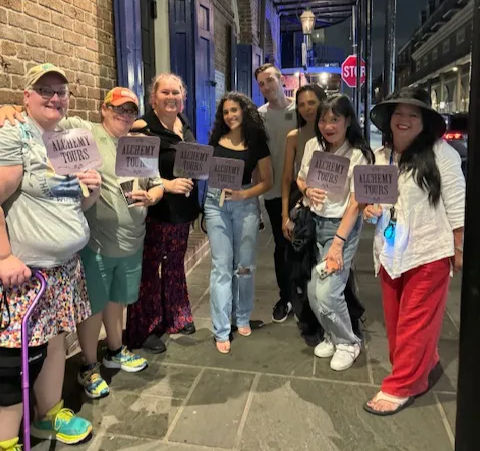 Smiling mixed-age walking-tour group posing on a nighttime historic-district sidewalk beneath iron balconies, each holding a small tour sign.