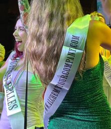 Women on stage wearing promotional sashes and party dresses — a green sequined dress with long hair and a laughing woman in white with beaded necklaces and a colorful sleeve tattoo, bathed in green stage lighting.