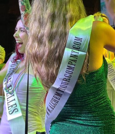 Women on stage wearing promotional sashes and party dresses — a green sequined dress with long hair and a laughing woman in white with beaded necklaces and a colorful sleeve tattoo, bathed in green stage lighting.