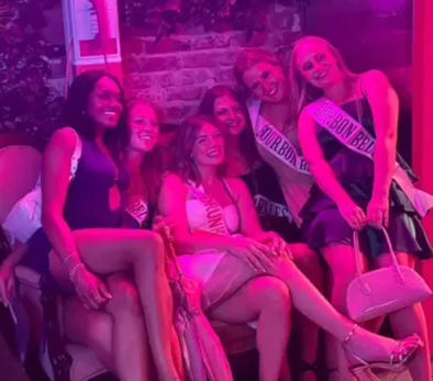 Six women in party dresses wearing sashes pose on a couch in a pink-lit bar lounge, smiling and holding small handbags against a brick wall backdrop.