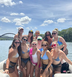 Smiling group of women in bikinis on a sunny boat party on a river with an arched bridge and blue skies, one wearing a pink sash.