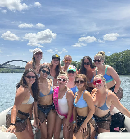 Smiling group of women in bikinis on a sunny boat party on a river with an arched bridge and blue skies, one wearing a pink sash.