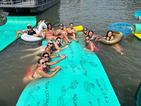 Group of women relaxing and laughing on teal floating foam pads and colorful pool inflatables near a dock on a sunny lake, holding drinks and enjoying a summer waterfront gathering