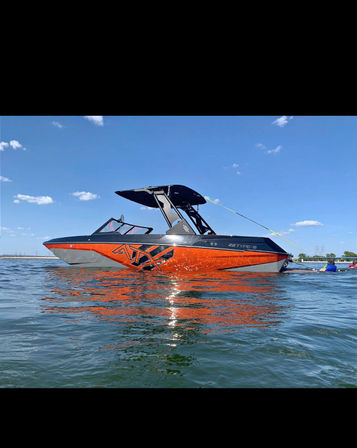 Orange and black wakeboat with a tall wakeboard tower and sunshade floating on a calm blue lake under a clear sunny sky, hull reflecting brightly in the water — ready for wakeboarding.