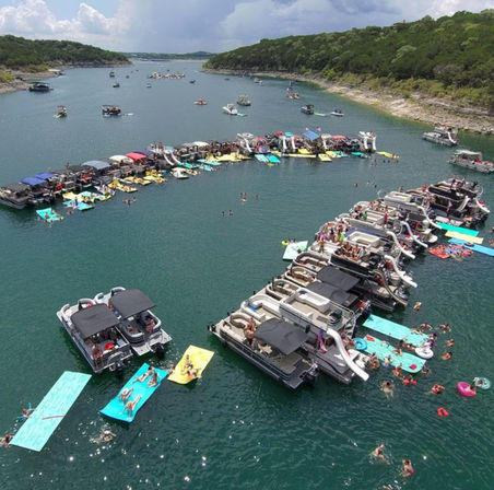 Aerial view of a lively summer lake party with dozens of pontoon boats anchored in a curved raft, colorful floating mats and slides, and swimmers in a tree-lined cove.