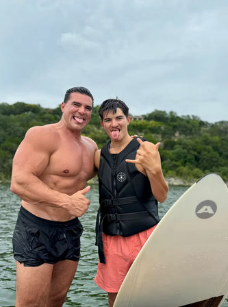 Two men standing in a lake beside a stand-up paddleboard — one shirtless giving a thumbs-up, the other in a black life vest and pink shorts flashing a shaka sign and sticking out his tongue, tree-lined shore and cloudy sky behind them.