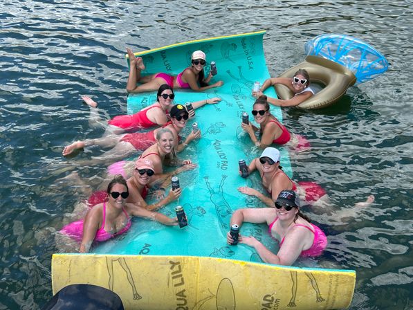 Group of friends in swimsuits lounging on a large turquoise floating mat in a lake, holding canned drinks next to a gold cocktail-shaped inflatable during a sunny summer float party.