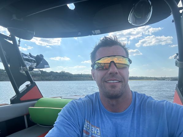 Smiling man on a motorboat wearing mirrored sports sunglasses and a blue shirt, with a sunlit lake, tree-lined shoreline and blue sky with scattered clouds in the background.