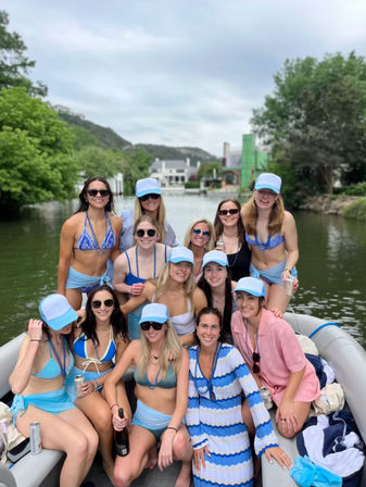 Group of women in blue bikinis and matching light-blue caps smiling together on a pontoon boat on a tree-lined river or lake, holding drinks and a champagne bottle on a cloudy summer day