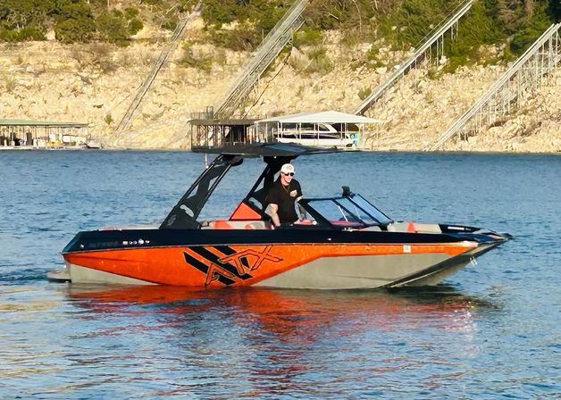 Orange-and-black wakeboard boat with a tower cruising on a blue lake near rocky shoreline and boat docks, person at the helm wearing a white cap