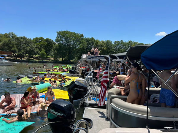 Crowded, festive summer lake party at a dock with pontoon boats, American flags, and people on bright inflatable mats and swimming under a clear blue sky