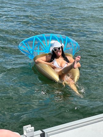Summer lake scene: woman in white bikini and straw hat with heart-shaped sunglasses lounging on a gold inflatable float with blue canopy, playfully sticking out her tongue near a boat