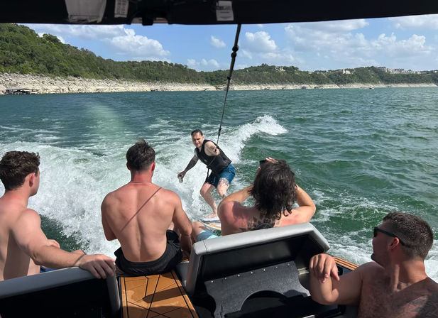 Group of friends on a motorboat watching a wakesurfer glide the boat wake on a sunny lake with tree-covered rocky shoreline and blue sky, summer water sports scene.