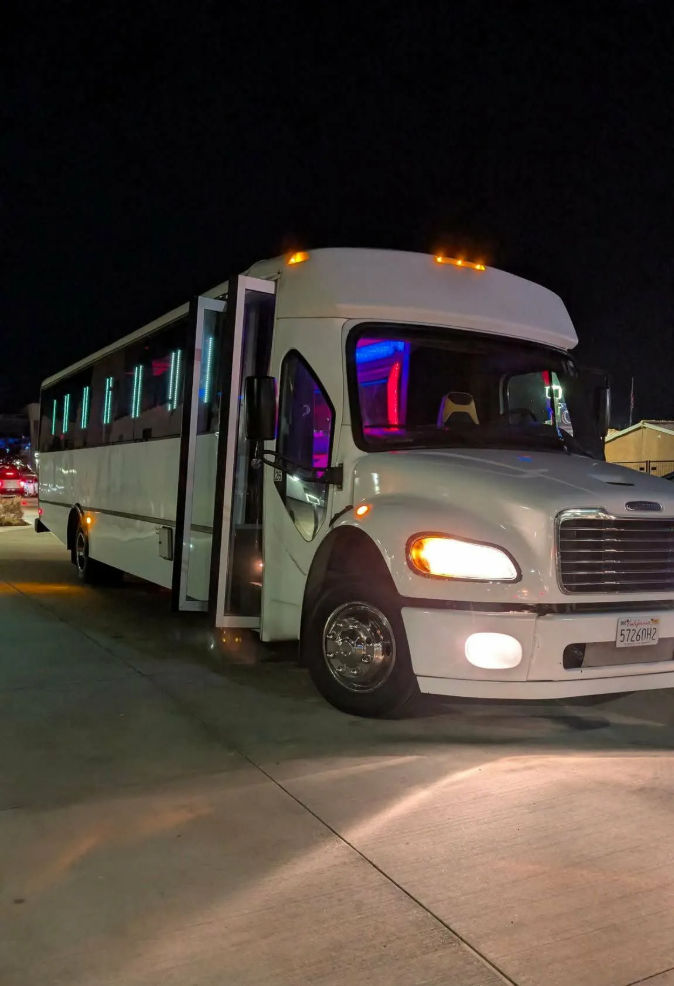 White party bus parked in a lot at night with its door open, headlights on and colorful LED neon interior lights glowing