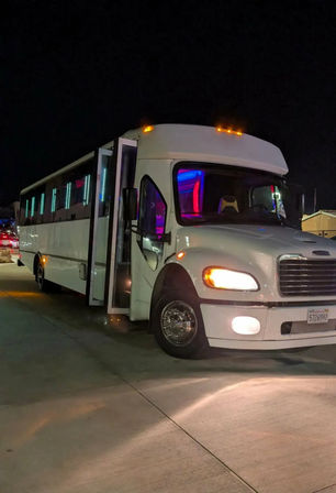 White party bus parked in a lot at night with its door open, headlights on and colorful LED neon interior lights glowing