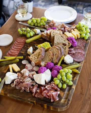 Wooden charcuterie board grazing platter with sliced artisan bread, assorted cured meats (salami, prosciutto), cheeses (brie, goat cheese, cheddar), green grapes, pickles and cornichons, radishes, bright purple pickled cauliflower, herb butter spread and wine glasses on a table.