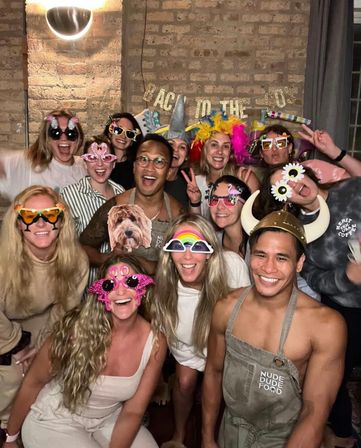 Smiling group of friends at an indoor brick-walled party wearing colorful novelty glasses, wigs, and a Viking helmet.