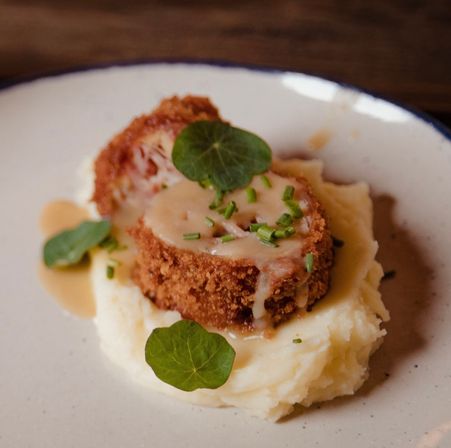 Crispy breaded meat medallion sliced and drizzled with creamy gravy, sprinkled with chopped chives and nasturtium leaves, served on a bed of mashed potatoes on a restaurant plate.