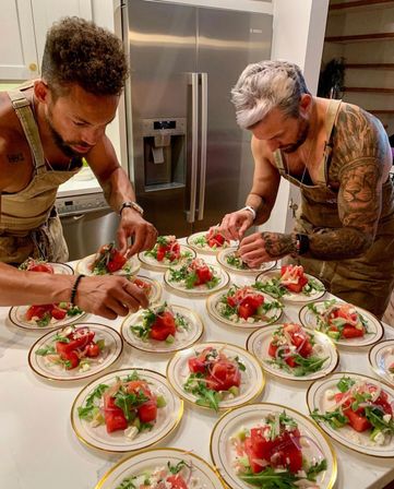 Two men in aprons carefully plating watermelon and arugula salads on gold-rimmed plates in a modern home kitchen with a stainless-steel fridge — dozens of elegant appetizer plates lined up for serving.