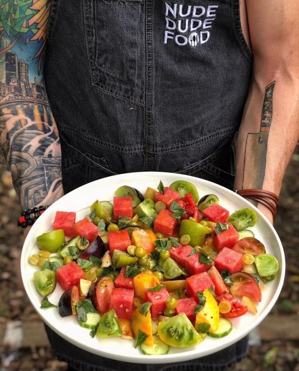 Tattooed arms holding a white plate of vibrant summer salad—cubed watermelon, mixed heirloom tomatoes, fresh basil and a light dressing
