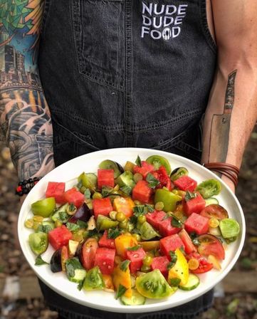 Tattooed arms holding a white plate of vibrant summer salad—cubed watermelon, mixed heirloom tomatoes, fresh basil and a light dressing