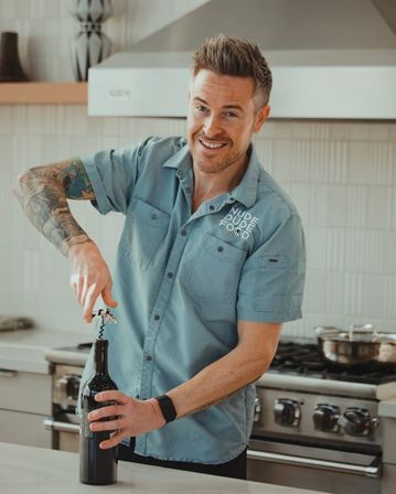 Smiling tattooed man using a corkscrew to open a wine bottle at a bright modern kitchen island with a stainless-steel range in the background.