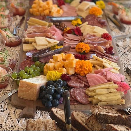 Rustic charcuterie board on a wooden farmhouse table with assorted cheeses, cured meats, green and black grapes, pickles, crusty bread and edible flowers.