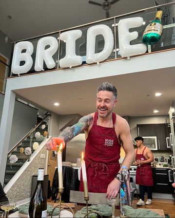 Smiling tattooed man in a red apron lights taper candles at a dining table in a modern kitchen, while large white "BRIDE" balloons and a champagne bottle balloon float along the upstairs railing.
