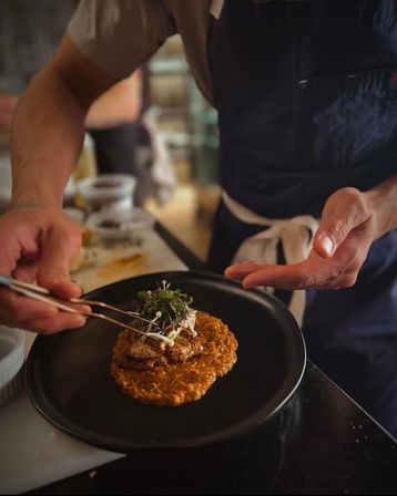 Hands-on chef plating a gourmet restaurant dish in a busy kitchen — crispy fritter on creamy tomato risotto, drizzled with sauce and topped with microgreens on a matte black plate.