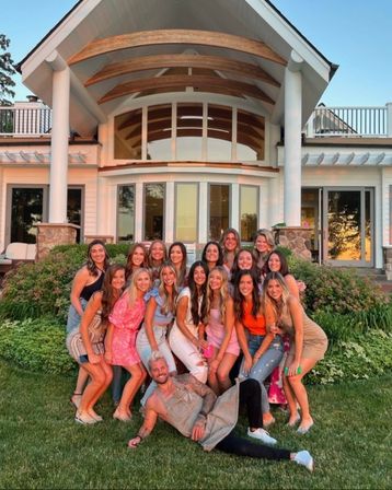Group of smiling young adults posing on a green lawn in front of a large modern house with arched wooden porch and big windows, bathed in warm sunset light — summer backyard friends photo.