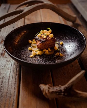 Gourmet seared pork belly cube topped with mango salsa and microgreens on cucumber rounds, served in a speckled black bowl on a rustic wooden table