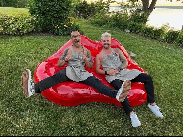 Two people in gray aprons laughing and posing on a large red lips-shaped inflatable sofa on a grassy lakeside lawn at sunset.