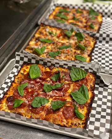 Rectangular Detroit-style pepperoni pizza with crispy caramelized crust, melted cheese, grated Parmesan and fresh basil on checkered-paper trays in a kitchen.