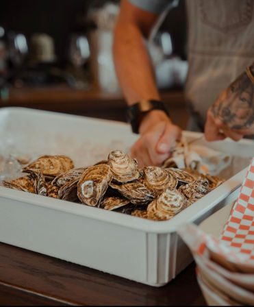Raw oysters piled on ice in a white bin at a seafood counter, hands in the background shucking shells