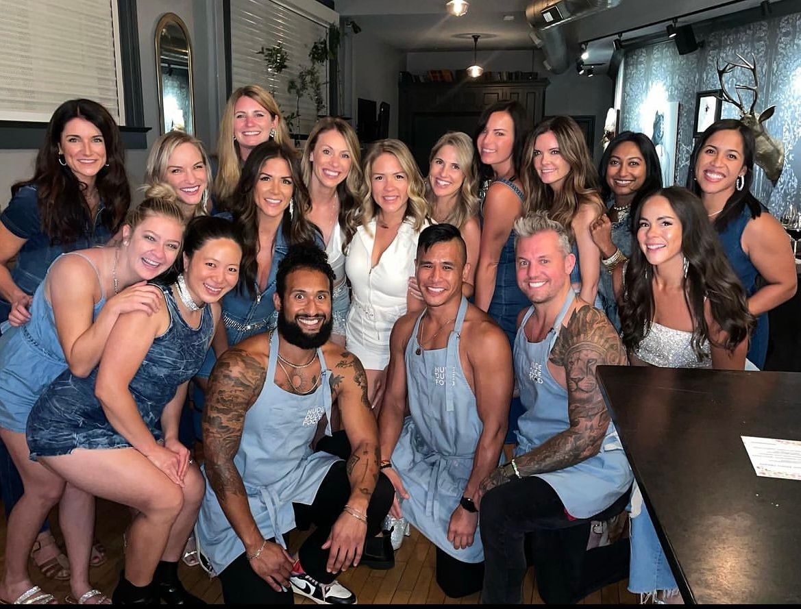 Group photo of about 19 smiling adults at a cozy indoor restaurant party, women in denim outfits and three apron-wearing men kneeling in front near a bar with decorative wall mount.