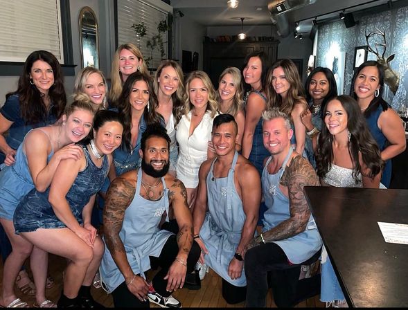 Group photo of about 19 smiling adults at a cozy indoor restaurant party, women in denim outfits and three apron-wearing men kneeling in front near a bar with decorative wall mount.