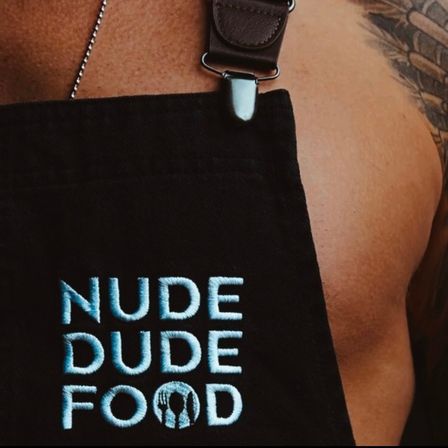 Close-up of a black cooking apron with light-blue embroidered 'NUDE DUDE FOOD' logo clipped by a leather strap over a bare-chested torso with a shoulder tattoo and thin chain necklace.