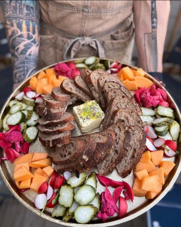 Artisan sourdough spiral with herbed butter centerpiece on a round grazing platter, surrounded by pickles, pink pickled cauliflower, radishes and cantaloupe.