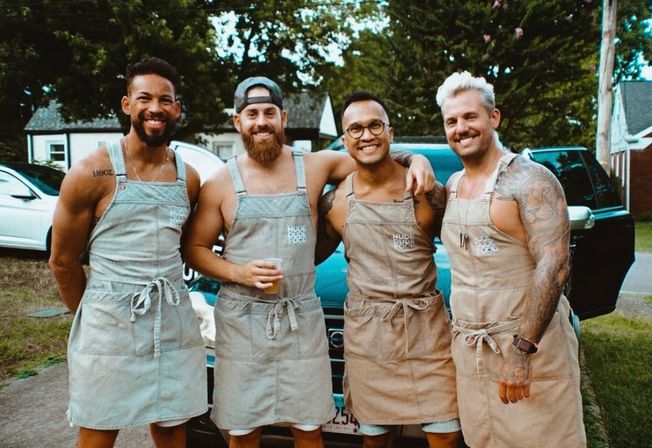 Four smiling men in matching canvas aprons posing arm-in-arm in front of a parked SUV on a suburban driveway, one holding a drink for a casual backyard cookout vibe.