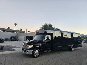 Sleek black party shuttle parked on a palm‑lined residential street at dusk with modern homes and mountains in the background