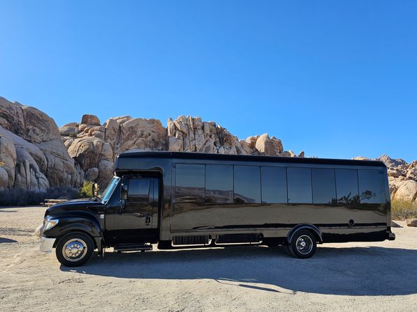 Black party bus parked on a sandy desert clearing with towering granite boulders and a vivid blue sky, the glossy side reflecting the rocky landscape.